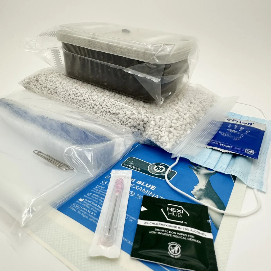 Show: The Grow Kit version of the XL Cylocybe Special MycBox seen from the side, its sealed tub of dark substrate resting on the bag of perlite, with the liner bag, nitrile gloves, face mask, alcohol and chlorhexidine wipes and a sterile syringe laid out in front